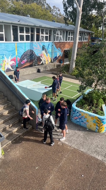 Students gathered around an outdoor table tennis table near a school building mural